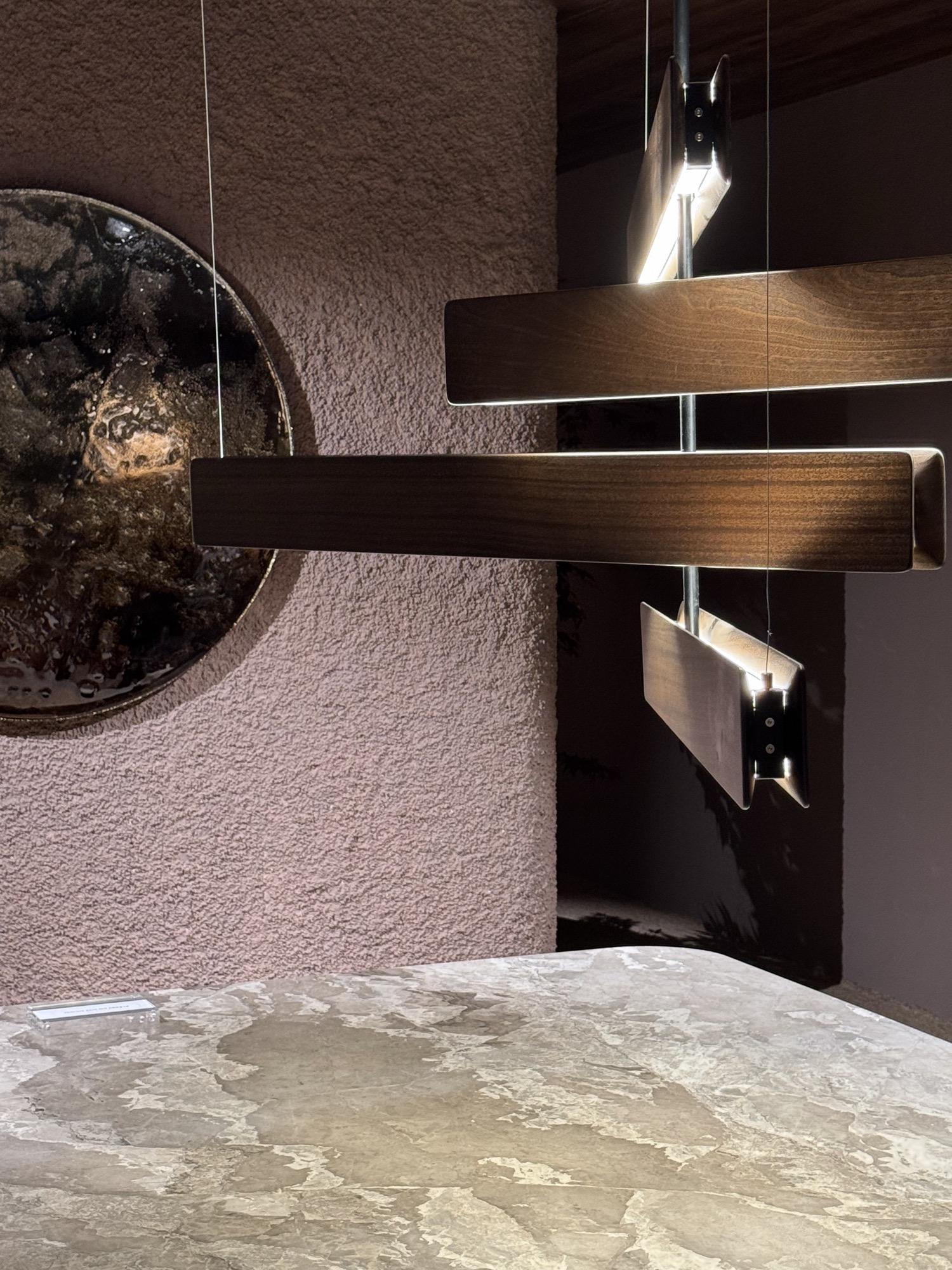 Suspended walnut light bars over a stone table against a pink plaster wall
