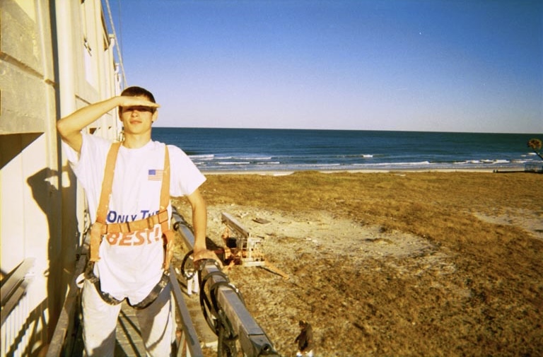 Young Roman in a safety harness on a facade platform, shading his eyes, the ocean behind